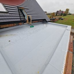 Fach plaski dach - Wykończony płaski dach z papy termozgrzewalnej, widoczne okna dachowe, ceglana attyka i narzędzia. W tle zabudowa jednorodzinna i pochmurne niebo.