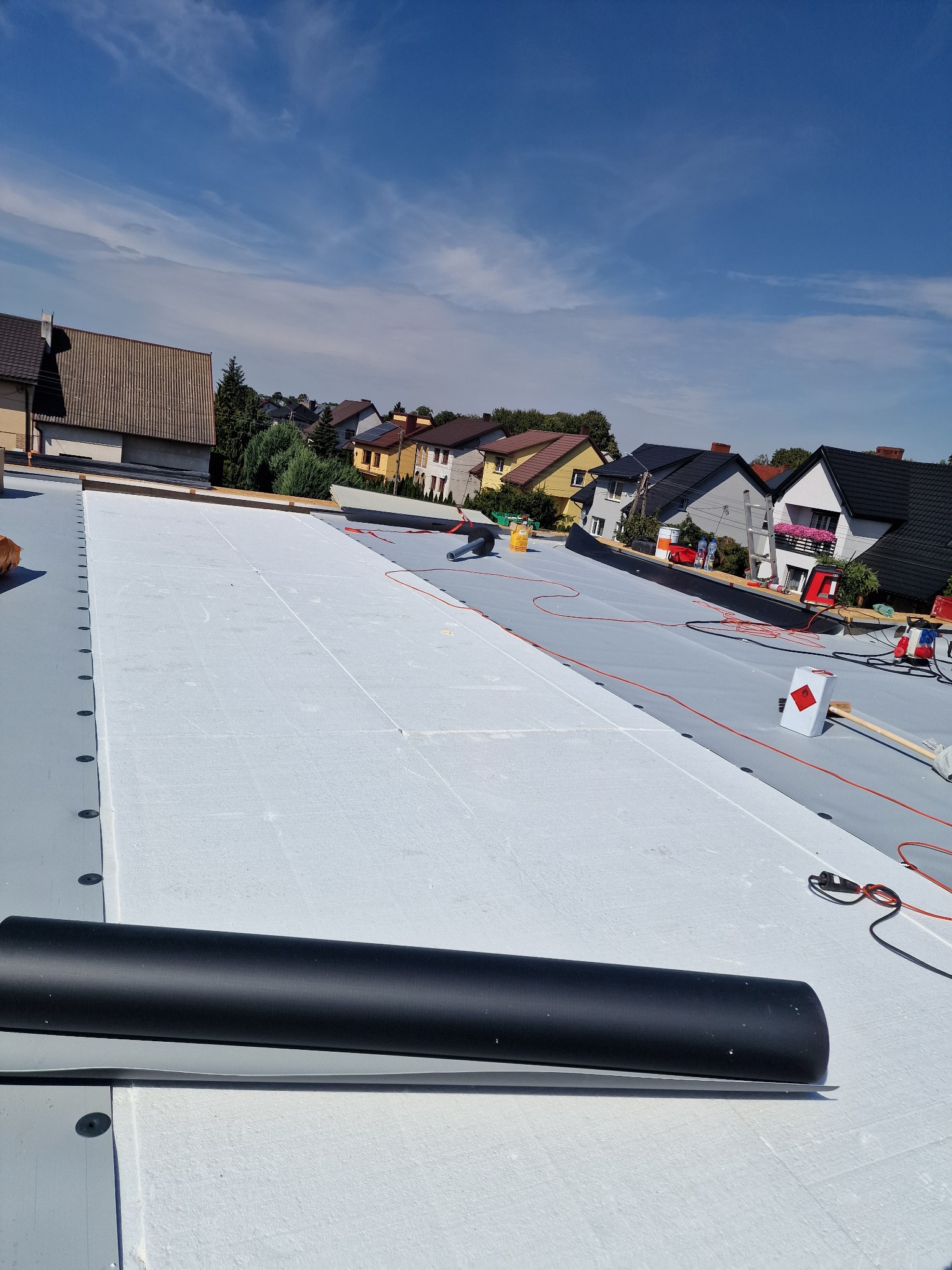 Biała termoizolacja na szarym dachu płaskim w trakcie montażu, widoczne narzędzia i rolka papy. W tle zabudowa jednorodzinna i błękitne niebo.