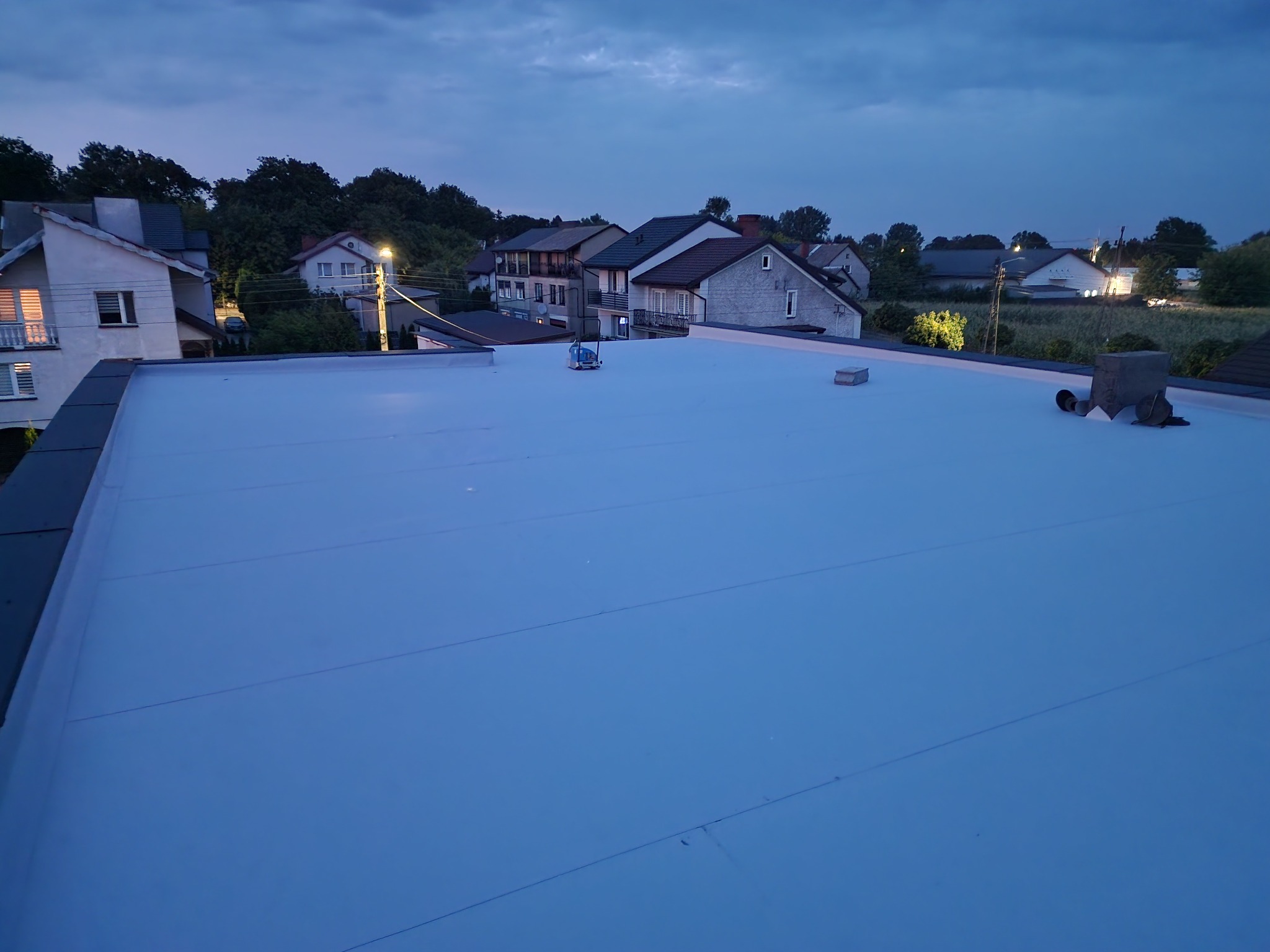 Nowy, płaski dach pokryty jasną membraną na tle zabudowań mieszkalnych o zmierzchu. Widok z góry ukazuje gładką powierzchnię i detale wykończenia.
