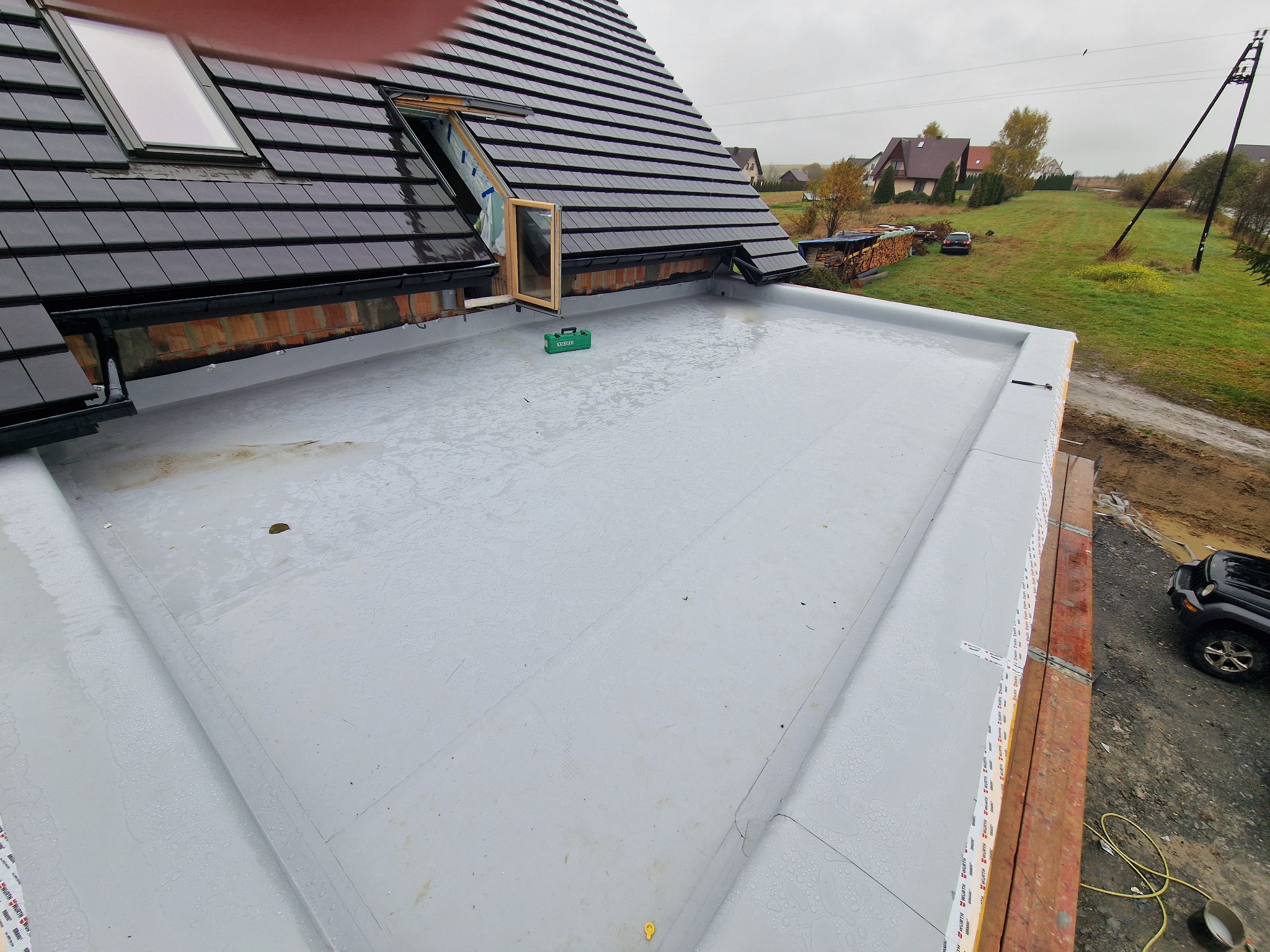 Wykończony płaski dach z papy termozgrzewalnej, widoczne okna dachowe, ceglana attyka i narzędzia. W tle zabudowa jednorodzinna i pochmurne niebo.