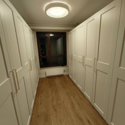 Pan Kanapa - Biała zabudowa szaf wnękowych z drewnianą podłogą i oknem. Złote uchwyty dodają elegancji. Oświetlenie sufitowe.