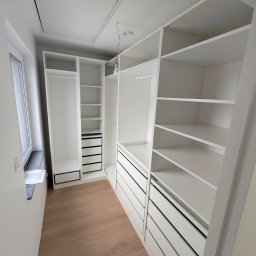 Pan Kanapa - Biała, nowoczesna garderoba z szufladami i półkami, widok z perspektywy wnętrza, drewniana podłoga, okno z lewej strony, minimalistyczny design.