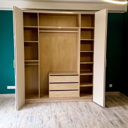 Pan Kanapa - Otwarta szafa wnękowa z jasnego drewna z szufladami i półkami, na tle zielonej ściany i drewnopodobnej podłogi. Widoczny nowoczesny żyrandol.