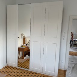 Pan Kanapa - Biała szafa z lustrem w sypialni, widoczna podłoga zabezpieczona kartonem, w tle pokój z biurkiem i rośliną. Widok na wnętrze przez otwarte drzwi.