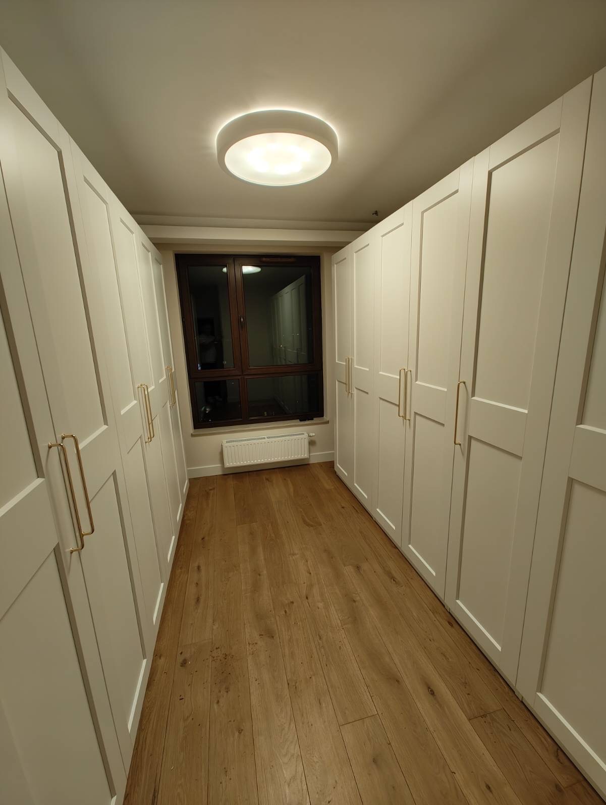 Biała zabudowa szaf wnękowych z drewnianą podłogą i oknem. Złote uchwyty dodają elegancji. Oświetlenie sufitowe.