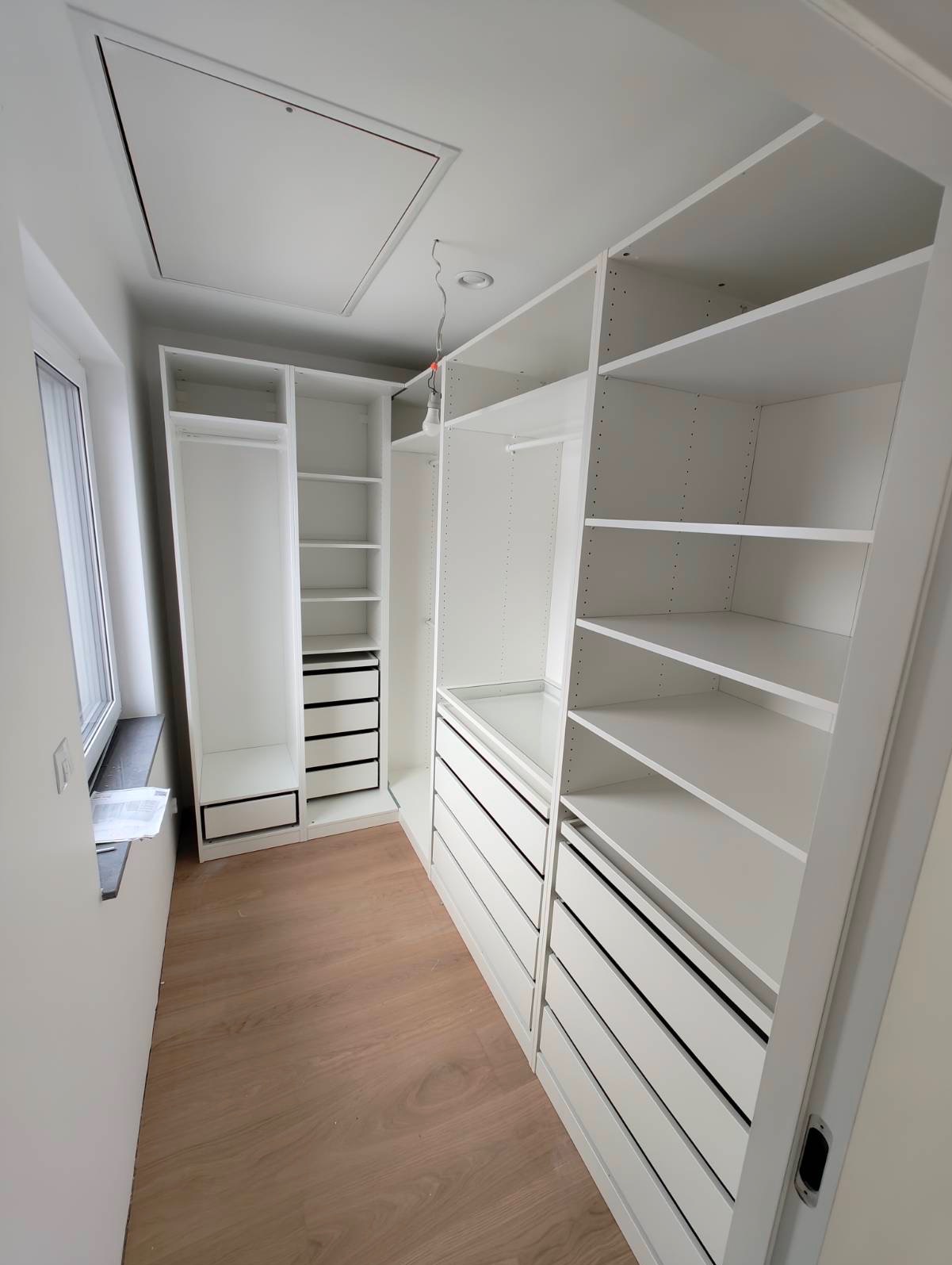 Biała, nowoczesna garderoba z szufladami i półkami, widok z perspektywy wnętrza, drewniana podłoga, okno z lewej strony, minimalistyczny design.