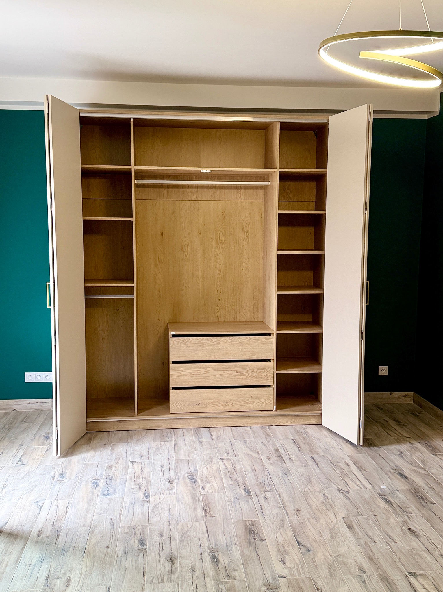 Otwarta szafa wnękowa z jasnego drewna z szufladami i półkami, na tle zielonej ściany i drewnopodobnej podłogi. Widoczny nowoczesny żyrandol.
