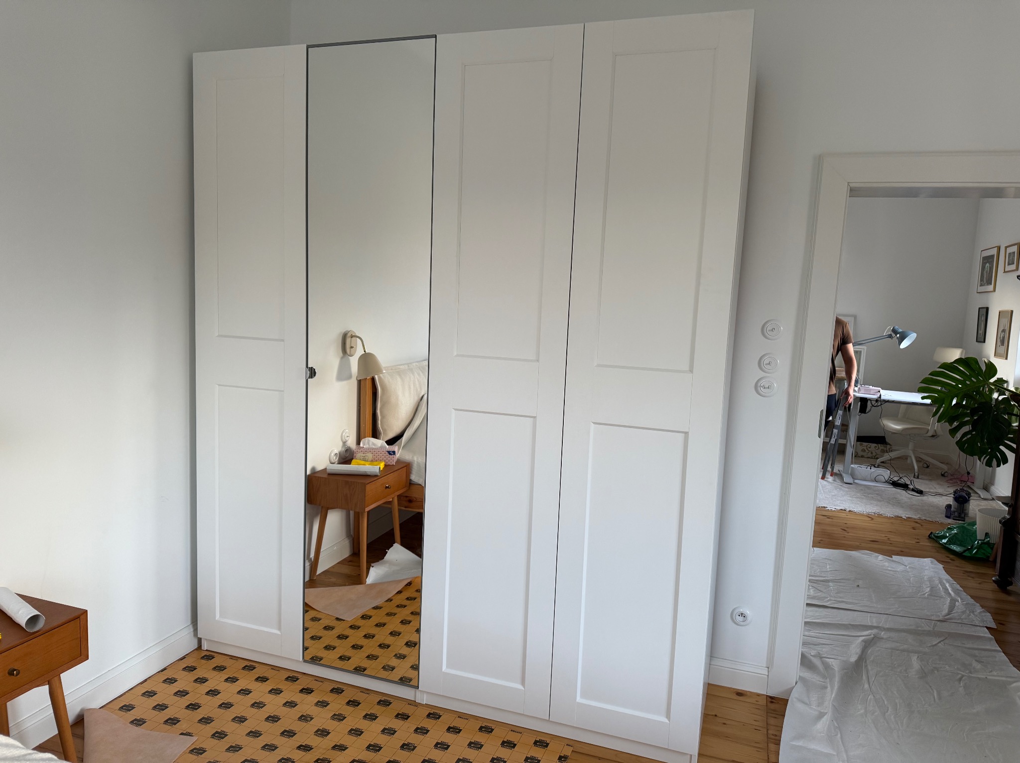 Biała szafa z lustrem w sypialni, widoczna podłoga zabezpieczona kartonem, w tle pokój z biurkiem i rośliną. Widok na wnętrze przez otwarte drzwi.