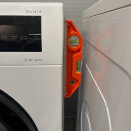 Yurii Herych - Poziomica między pralką a suszarką, sprawdzanie wypoziomowania. Białe urządzenia AGD w łazience z szarymi płytkami w tle. Pomarańczowy przyrząd pomiarowy.