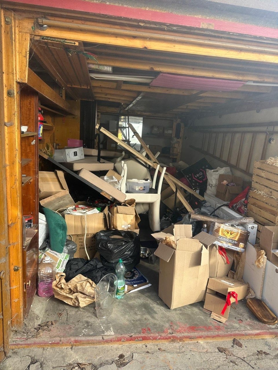 Zdemolowane wnętrze z chaotycznym nagromadzeniem mebli, kartonów i odpadów. Widoczne elementy konstrukcyjne budynku. Scena po wyburzeniu lub w trakcie przeprowadzki.