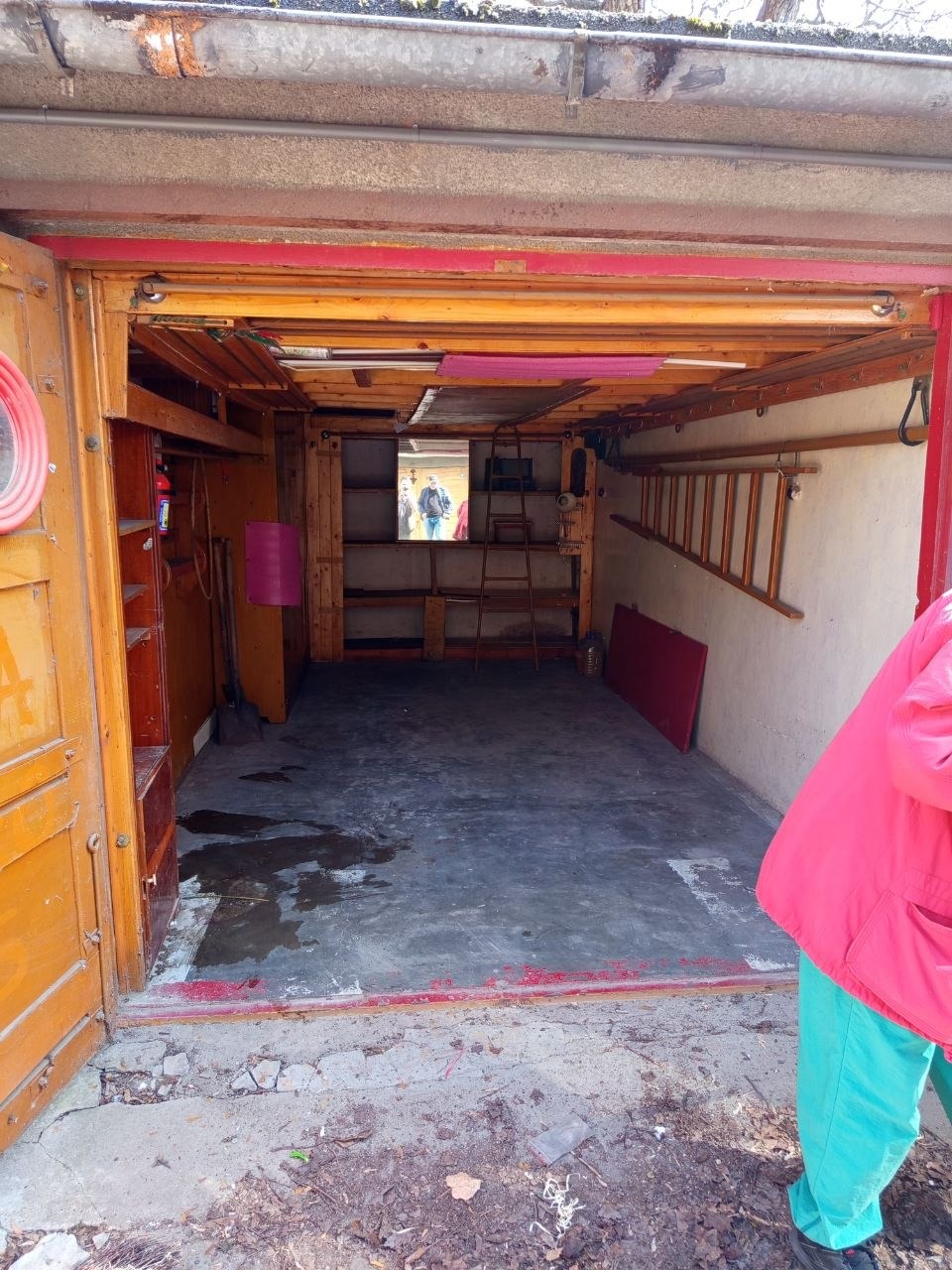 Wnętrze garażu z drewnianą konstrukcją, widoczne półki, drabina i betonowa podłoga. Częściowo otwarte, pomalowane na żółto drzwi garażowe.