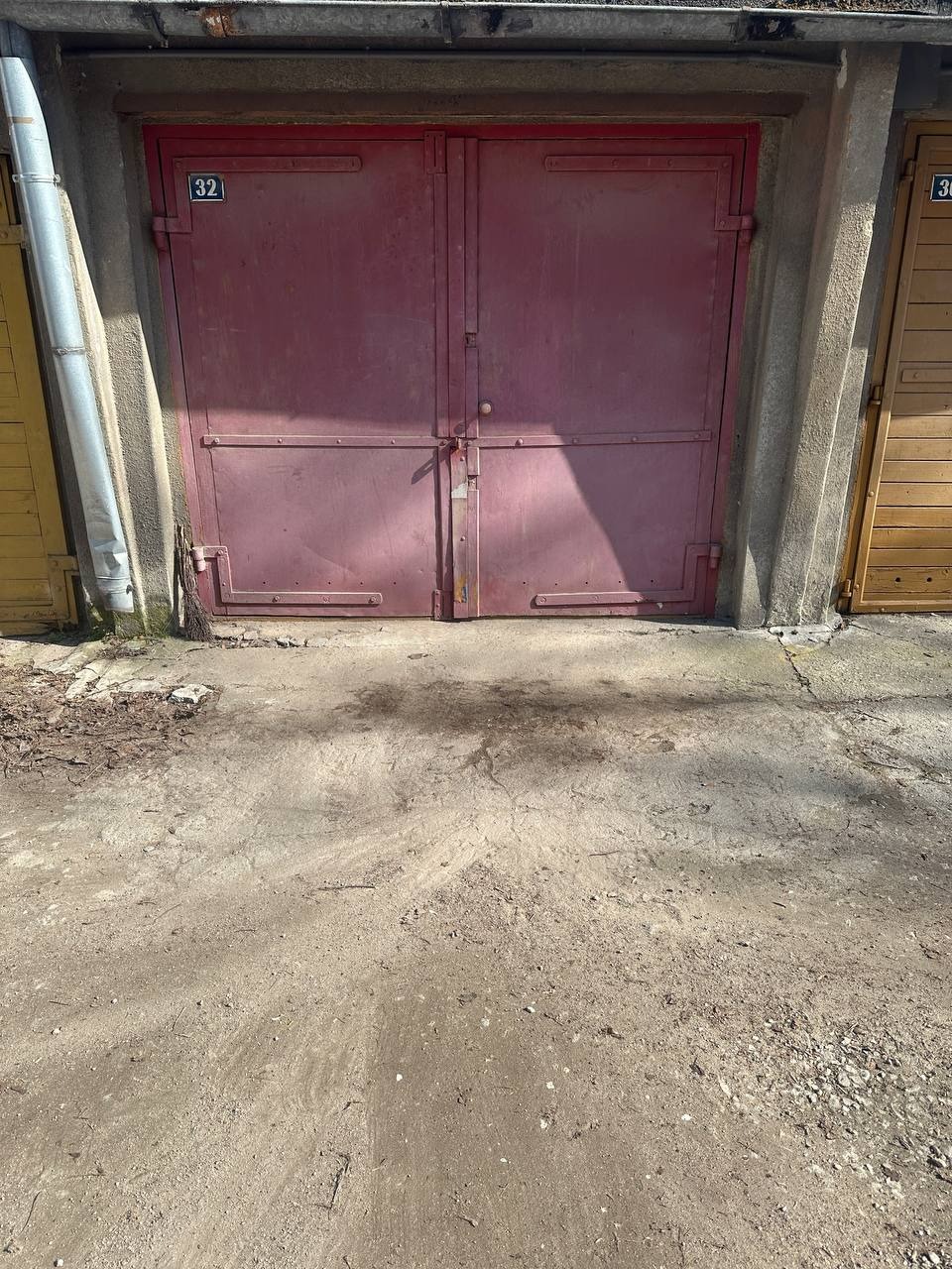 Czerwone, metalowe drzwi garażowe z numerem 32, osadzone w betonowej konstrukcji. Obok widoczne fragmenty żółtych i drewnianych drzwi garażowych z numerem 36.