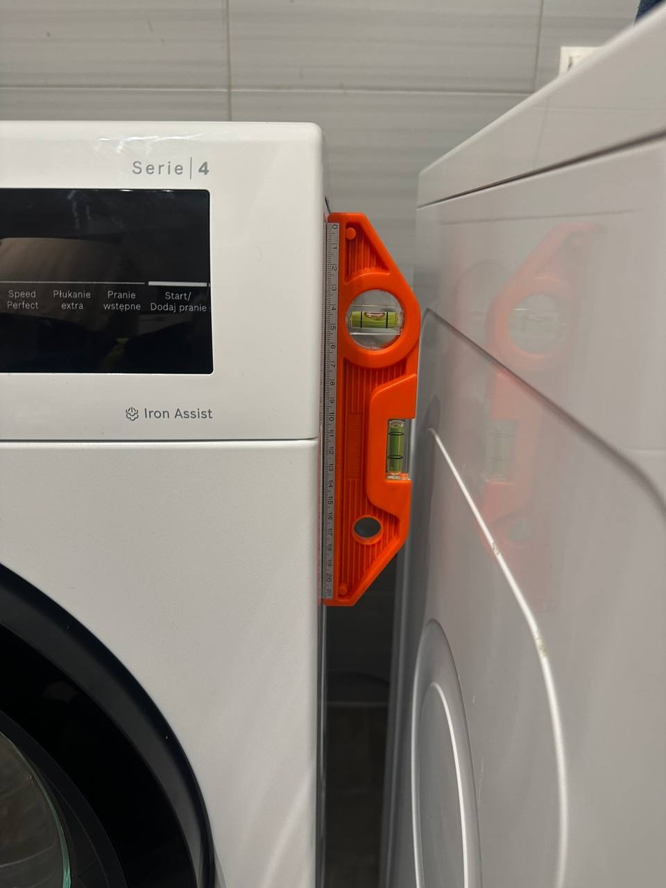 Poziomica między pralką a suszarką, sprawdzanie wypoziomowania. Białe urządzenia AGD w łazience z szarymi płytkami w tle. Pomarańczowy przyrząd pomiarowy.