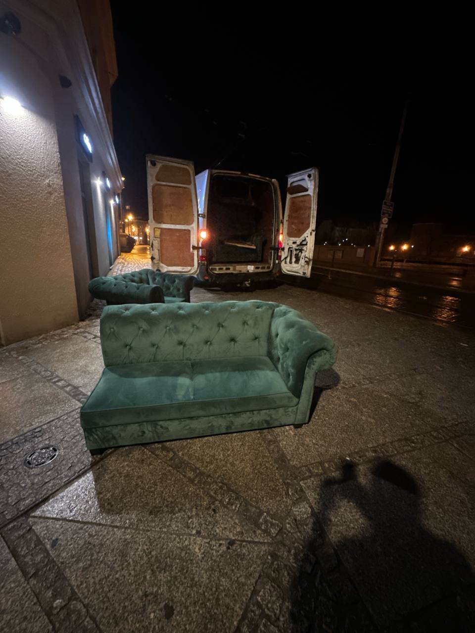 Turkusowa sofa chesterfield na chodniku, otwarte drzwi busa w tle. Nocna sceneria, mokra nawierzchnia. Nietypowe ujęcie mebla przed transportem.