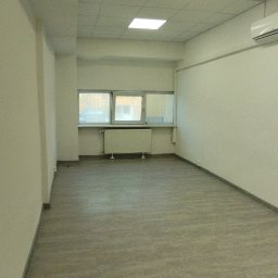 Ivan Rus - Wykończone wnętrze biurowe z szarą podłogą, białymi ścianami i sufitem kasetonowym. Widoczne okna z grzejnikiem i klimatyzator. Minimalistyczny, nowoczesny design.