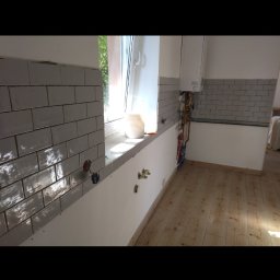 Ivan Rus - Kuchnia w trakcie wykończenia: szare płytki ułożone na ścianie, widoczne instalacje wodne i elektryczne, drewniana podłoga. Częściowo wykończone wnętrze.