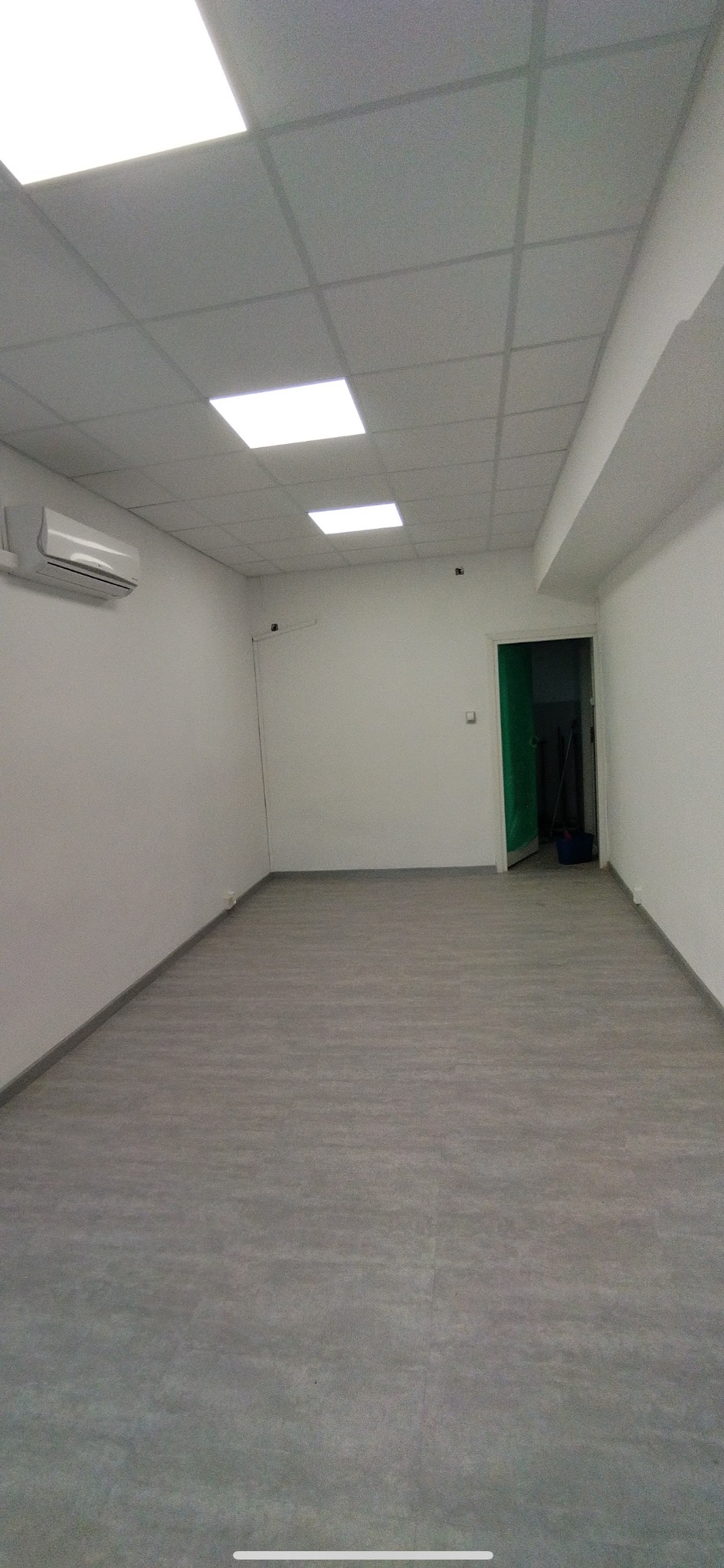 Wnętrze wyremontowanego biura z szarą podłogą, białymi ścianami i sufitem kasetonowym z oświetleniem LED. Widoczny klimatyzator i otwarte drzwi do innego pomieszczenia.