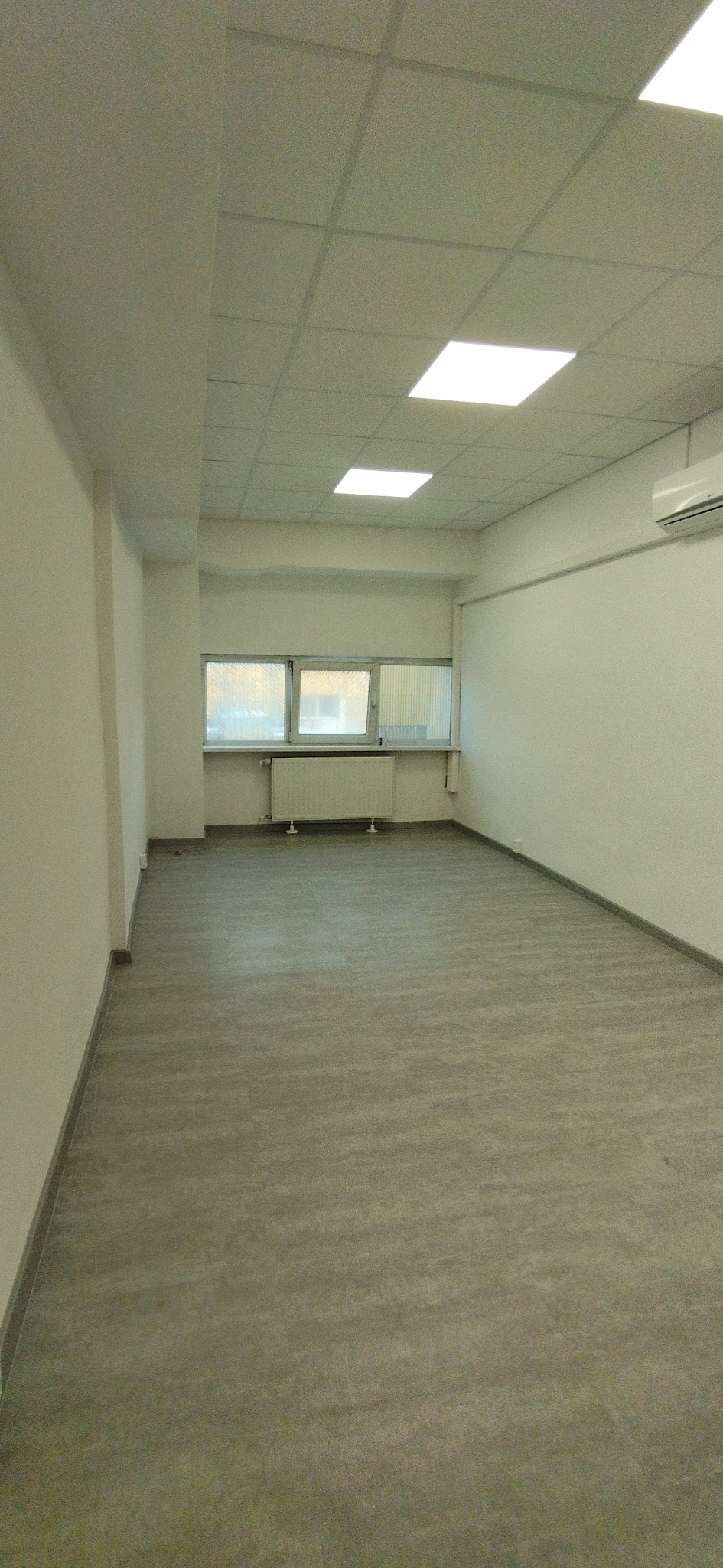 Wykończone wnętrze biurowe z szarą podłogą, białymi ścianami i sufitem kasetonowym. Widoczne okna z grzejnikiem i klimatyzator. Minimalistyczny, nowoczesny design.