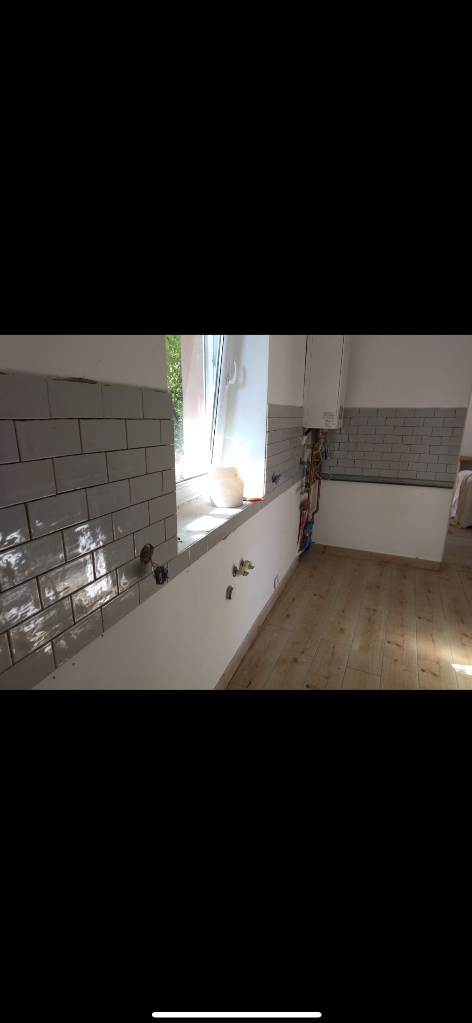 Kuchnia w trakcie wykończenia: szare płytki ułożone na ścianie, widoczne instalacje wodne i elektryczne, drewniana podłoga. Częściowo wykończone wnętrze.