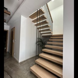 Adrian Węgrecki - Nowoczesne schody drewniane z czarną balustradą w jasnym wnętrzu. Widoczne detale wykończenia podłogi i minimalistyczny design.