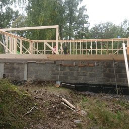 Adrian Węgrecki - Wznoszenie drewnianej konstrukcji domu jednorodzinnego na fundamencie z bloczków betonowych. Widoczne narzędzia i osoby pracujące na budowie.