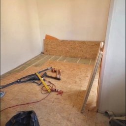 Adrian Węgrecki - Montaż podłogi z płyt OSB w pomieszczeniu. Widoczne narzędzia, poziomica, klej, płyty OSB o różnej wielkości. Częściowo ułożona podłoga na legarach.