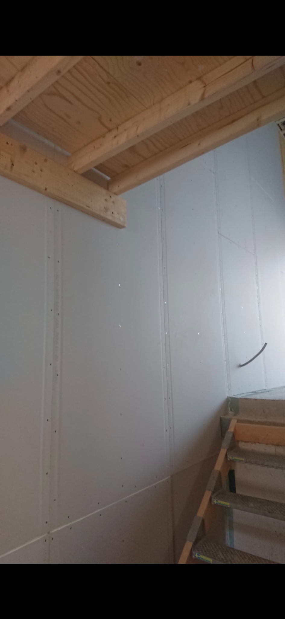 Wnętrze w trakcie prac wykończeniowych: ściany z płyt gipsowo-kartonowych, drewniana konstrukcja sufitu i fragment schodów. Widoczne łączenia płyt i ślady po wkrętach.