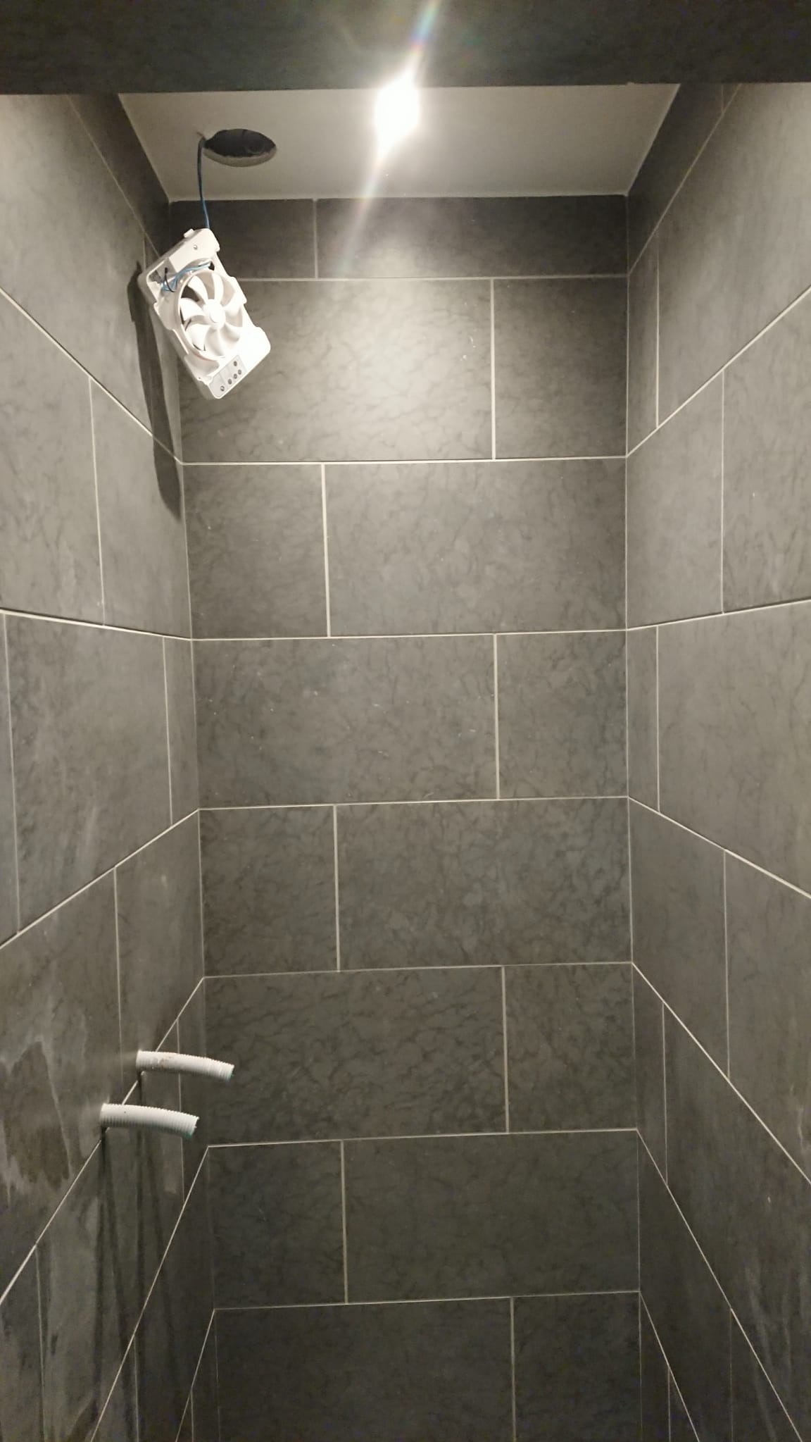 Szare płytki w kabinie prysznicowej z widocznymi rurami i wentylatorem w trakcie montażu. Oświetlenie sufitowe.