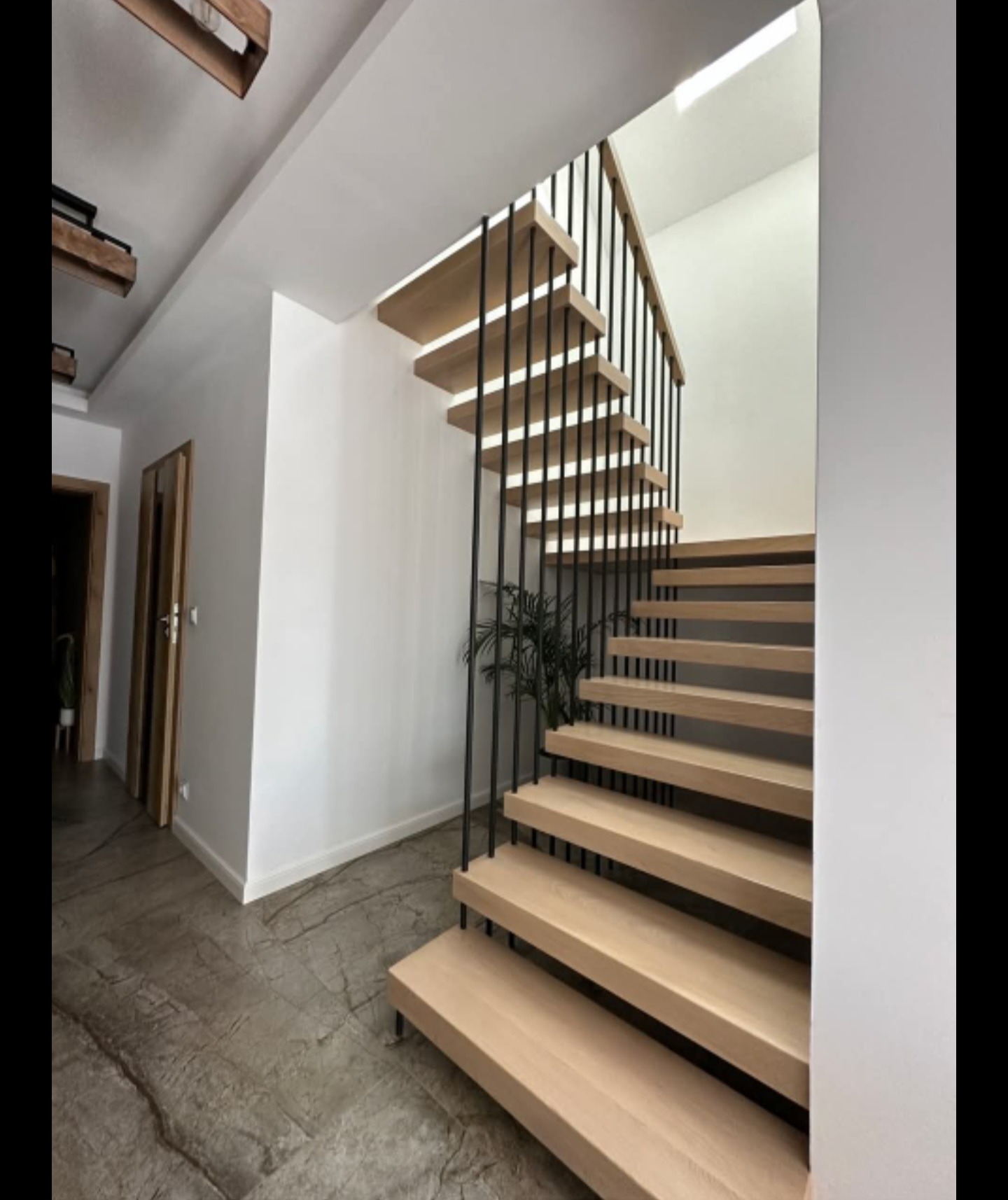 Nowoczesne schody drewniane z czarną balustradą w jasnym wnętrzu. Widoczne detale wykończenia podłogi i minimalistyczny design.