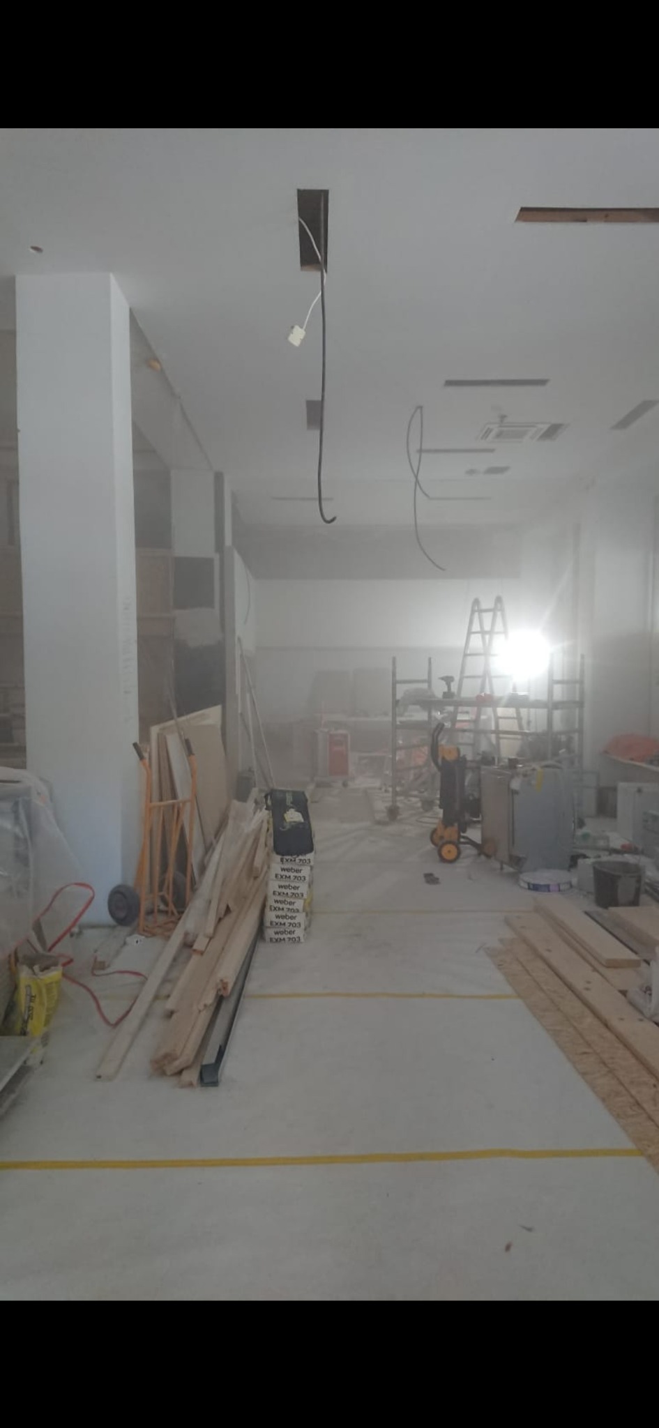 Wnętrze w trakcie prac wykończeniowych: widoczne materiały budowlane, drabina, instalacje elektryczne w suficie. W tle regały i narzędzia.