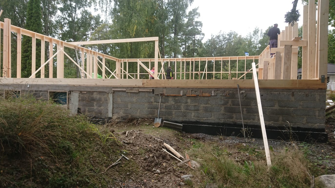 Wznoszenie drewnianej konstrukcji domu jednorodzinnego na fundamencie z bloczków betonowych. Widoczne narzędzia i osoby pracujące na budowie.