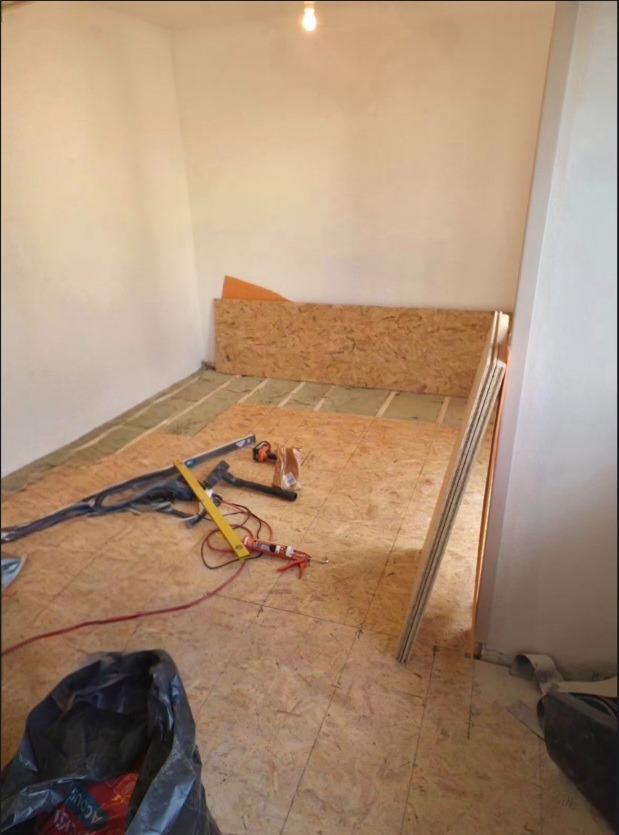 Montaż podłogi z płyt OSB w pomieszczeniu. Widoczne narzędzia, poziomica, klej, płyty OSB o różnej wielkości. Częściowo ułożona podłoga na legarach.