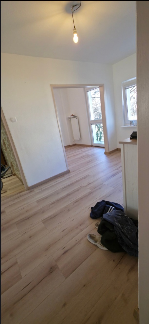 Jasne panele podłogowe w odnowionym pokoju z białymi ścianami i widokiem na okno. Uwagę zwraca wisząca żarówka bez klosza i ubrania leżące na podłodze.