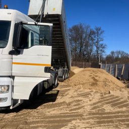 HANDEL - USŁUGI GRAŻYNA KOCHAN - Ciężarówka MAN TGX 18.440 z otwartą naczepą wysypuje piasek na plac budowy, widoczne ślady opon na ziemi i fragment ogrodzenia. Błękitne niebo w tle.