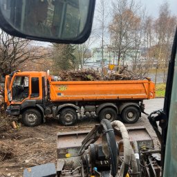 HANDEL - USŁUGI GRAŻYNA KOCHAN - Pomarańczowa wywrotka Meiller-Kipper załadowana gałęziami w Jeleniej Górze, widziana z kabiny innego pojazdu budowlanego. Prace ziemne w tle.