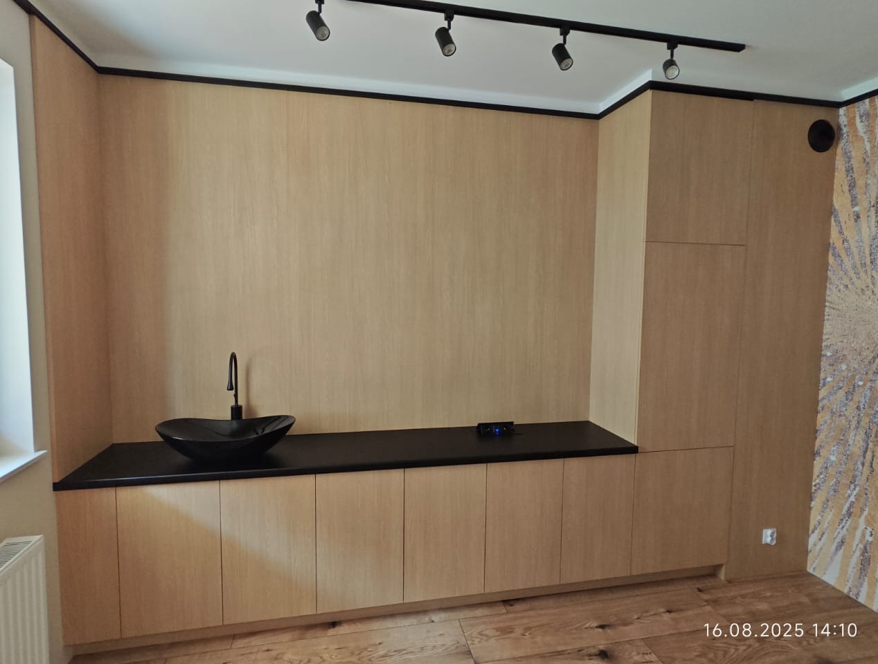 Jasne, drewniane szafki łazienkowe z czarnym blatem i umywalką w kształcie łzy. Minimalistyczne oświetlenie punktowe i drewniana podłoga.