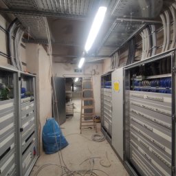 AK Elektro - Rozbudowana instalacja elektryczna w serwerowni: szafy rack, kable w korytach, drabina. Widoczne kable i złącza. Oświetlenie jarzeniowe. Etap prac w toku.
