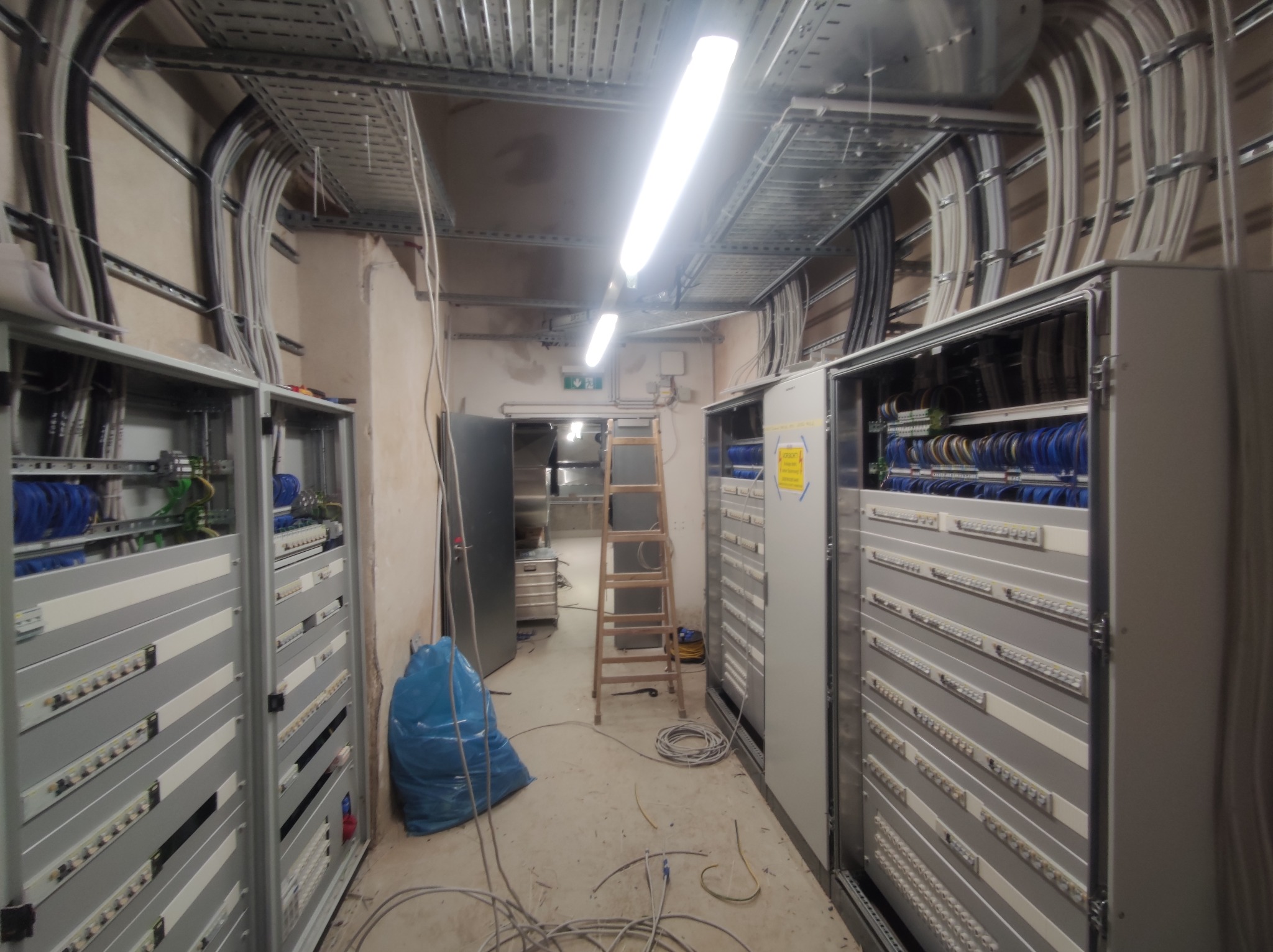 Rozbudowana instalacja elektryczna w serwerowni: szafy rack, kable w korytach, drabina. Widoczne kable i złącza. Oświetlenie jarzeniowe. Etap prac w toku.