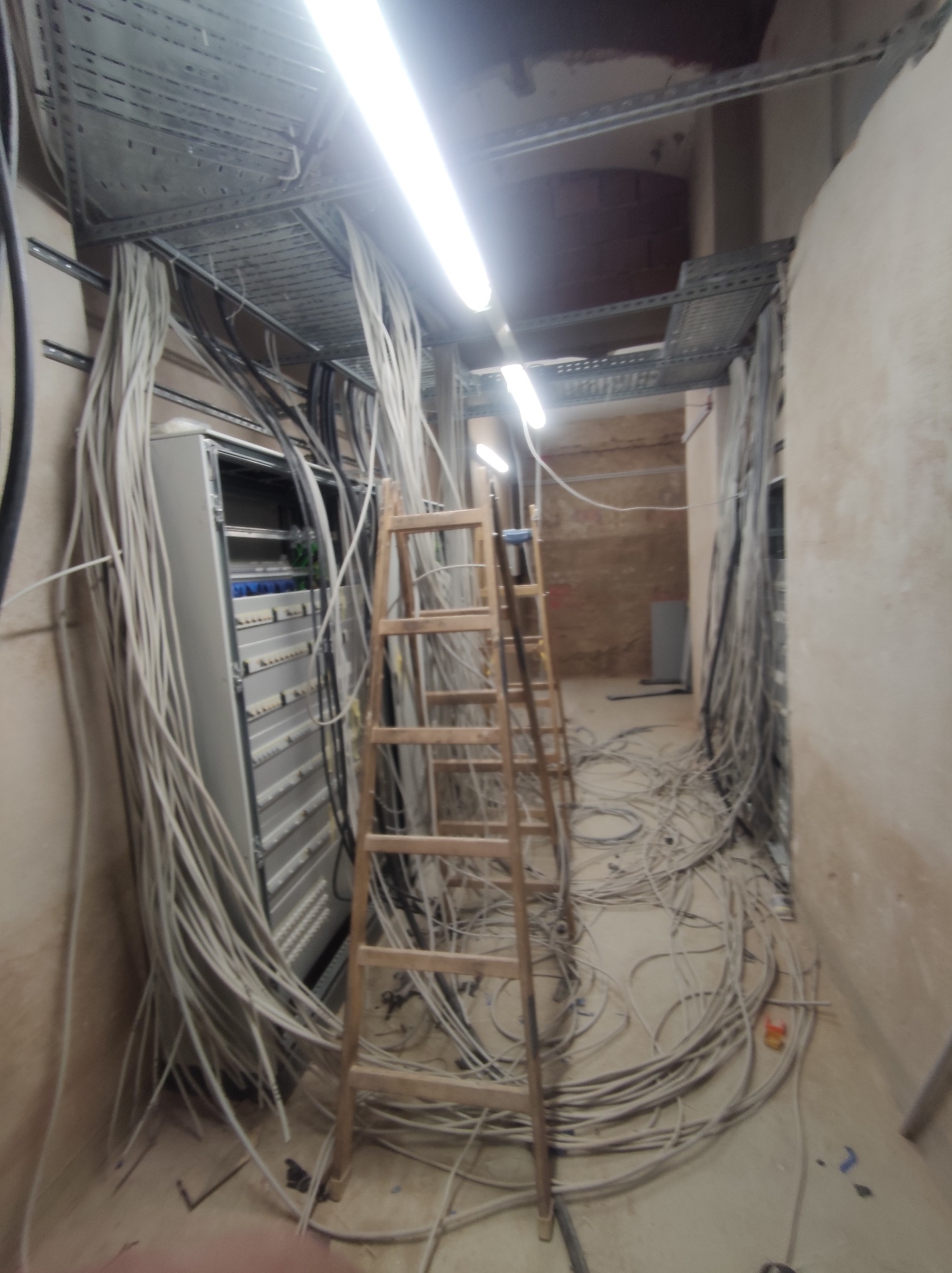 Rozbudowana instalacja elektryczna w budynku w trakcie prac. Widoczne kable, drabina i szafa rozdzielcza. Przewody ułożone na metalowych korytkach pod sufitem.