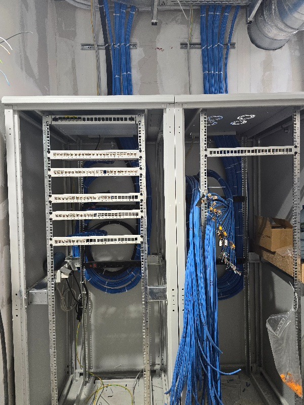 Szafa rack z niebieskimi kablami sieciowymi, widoczne kable ułożone wewnątrz i nad szafą, surowy stan wykończenia ścian, instalacja w trakcie realizacji.