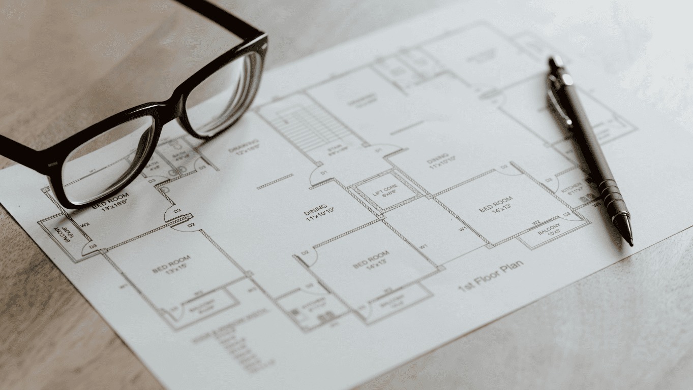 Plan architektoniczny z naniesionymi wymiarami pomieszczeń, czarny długopis i okulary na jasnym tle. Widok z góry, detale projektu.