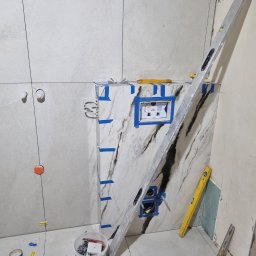 Układanie płytek w łazience w Wągrowcu. Widoczny poziomica, płytki imitujące marmur, oraz narzędzia glazurnicze. Prace w toku, surowy stan.