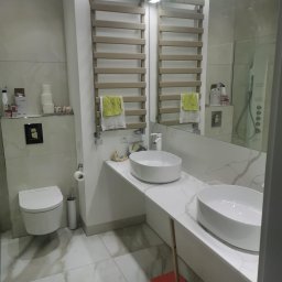 Nowoczesna łazienka z marmurową posadzką i blatem, dwie białe umywalki nablatowe, grzejnik drabinkowy z ręcznikiem i toaleta podwieszana. Minimalistyczny design.