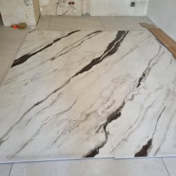 Ułożone, marmurowe płytki podłogowe z ciemnymi żyłami, kontrastujące z surowym tłem ściany w trakcie prac. Widoczne detale instalacji elektrycznej.