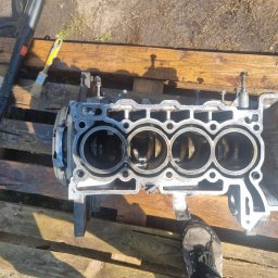 PAW-BUD - Czyszczenie bloku silnika spalinowego na drewnianej palecie, widoczna myjka ciśnieniowa i pędzel. Blok czterocylindrowy, z widocznymi tłokami. Noga w bucie w prawym dolnym rogu.