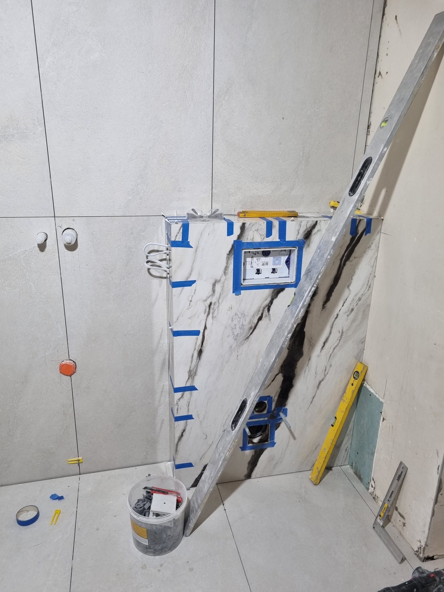 Układanie płytek w łazience w Wągrowcu. Widoczny poziomica, płytki imitujące marmur, oraz narzędzia glazurnicze. Prace w toku, surowy stan.