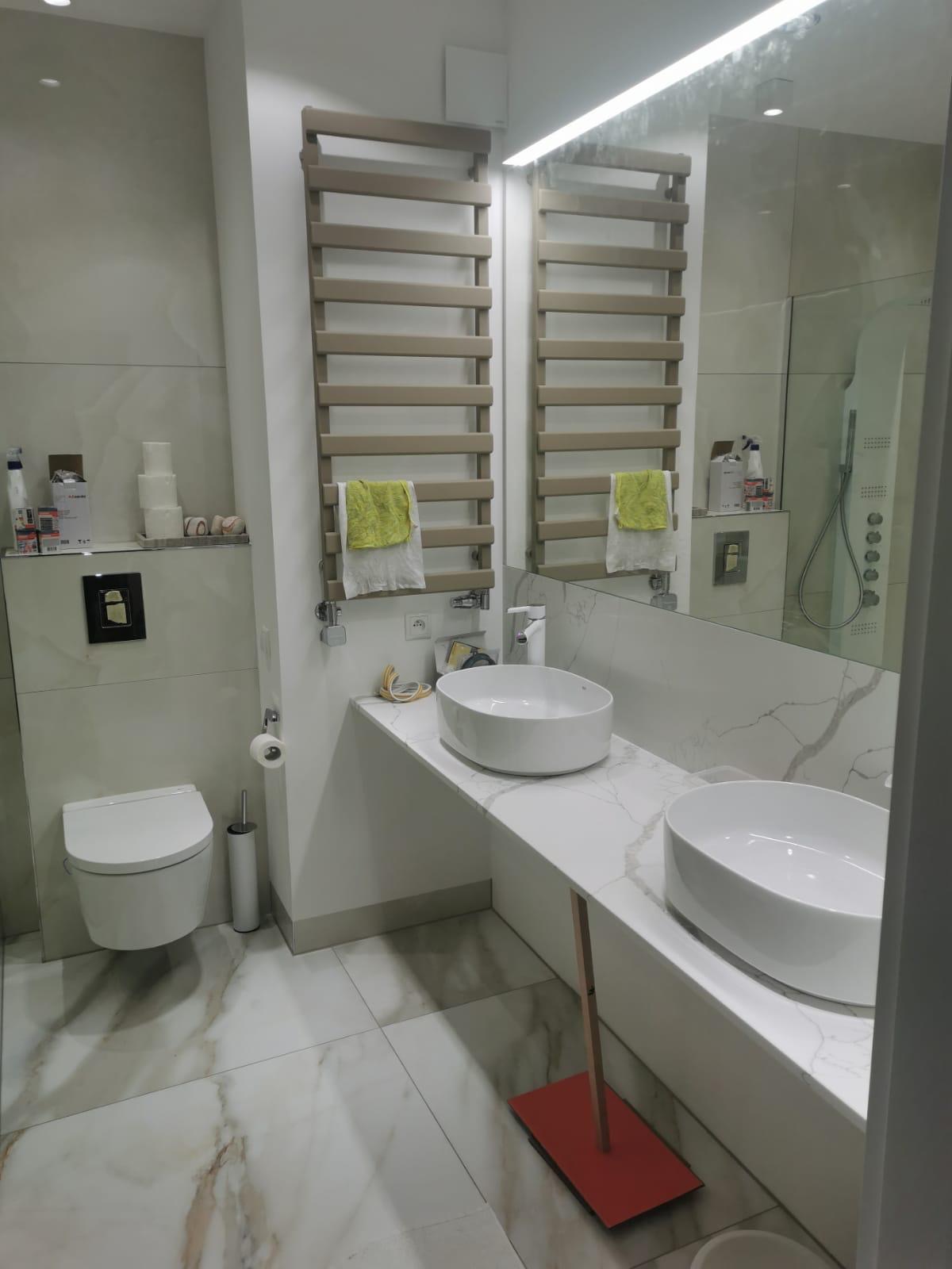 Nowoczesna łazienka z marmurową posadzką i blatem, dwie białe umywalki nablatowe, grzejnik drabinkowy z ręcznikiem i toaleta podwieszana. Minimalistyczny design.