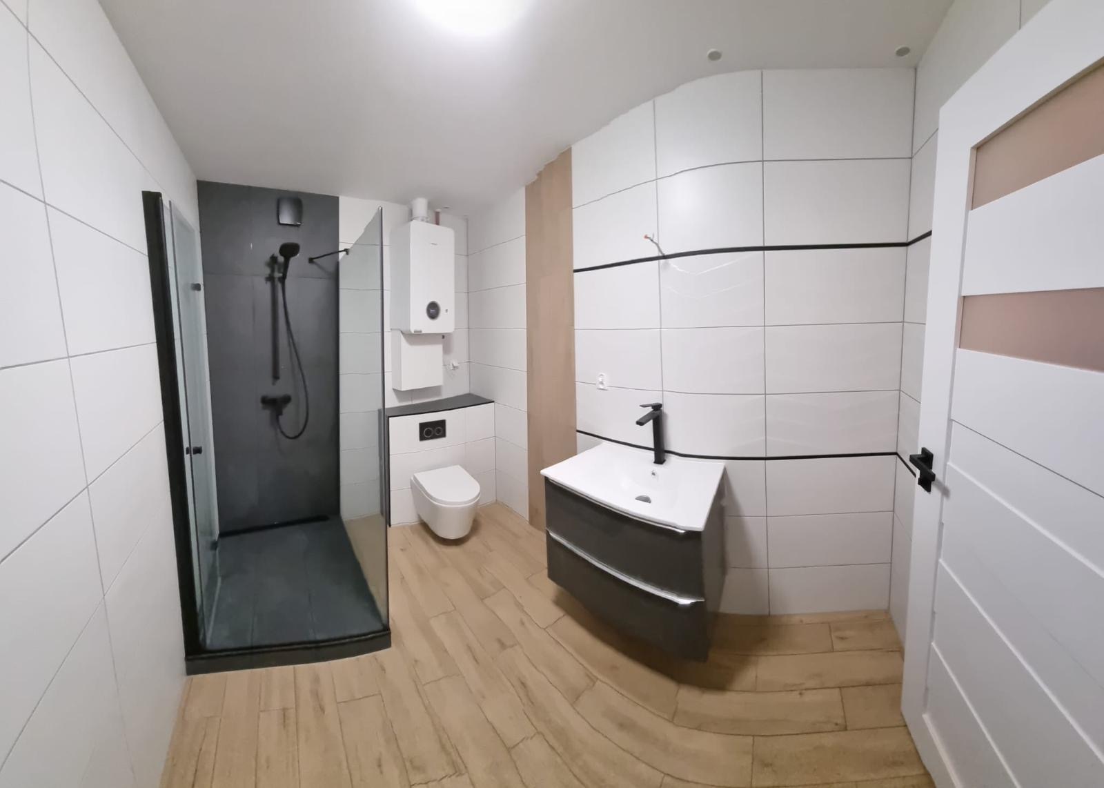 Nowoczesna łazienka z kabiną prysznicową typu walk-in, umywalką na szafce i podwieszaną toaletą. Białe płytki z czarnym akcentem i drewnopodobna podłoga.