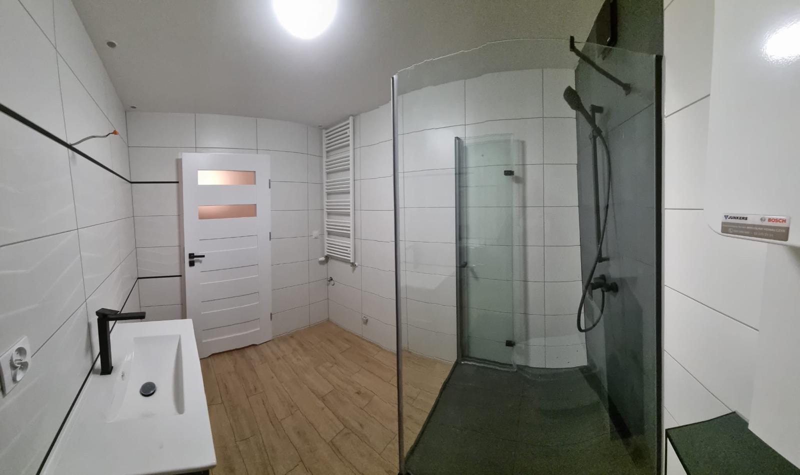 Nowoczesna łazienka po remoncie: białe płytki, kabina prysznicowa ze szkła, drewnopodobna podłoga, biała umywalka z czarną armaturą i grzejnik drabinkowy.