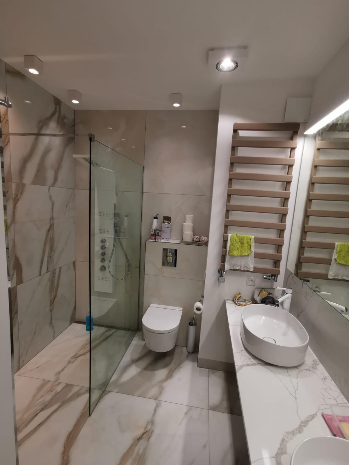Nowoczesna łazienka z kabiną prysznicową typu walk-in, umywalką nablatową i grzejnikiem drabinkowym. Marmurowe płytki na ścianach i podłodze. Oświetlenie punktowe.